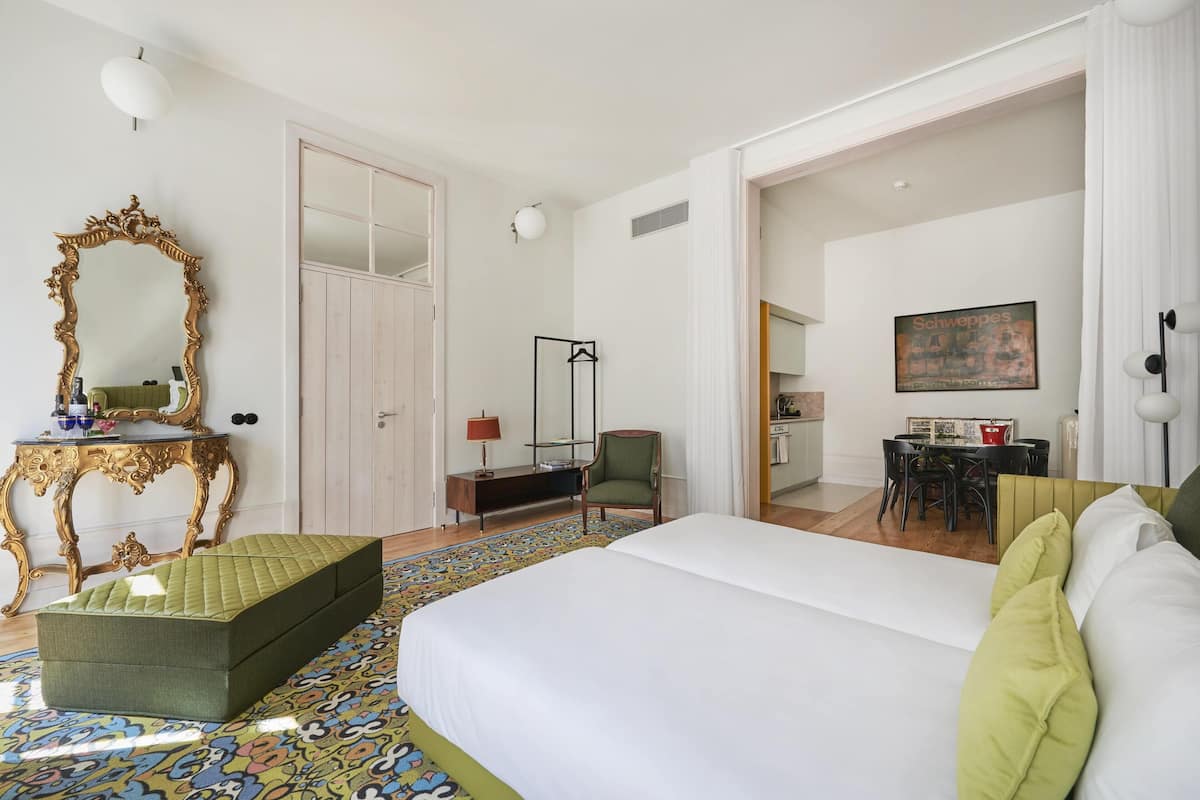 Habitación de hotel con una cama grande, un reposapiés verde, un espejo con marco ornamentado, un escritorio con lámpara y un área de comedor visible a través de una puerta abierta.