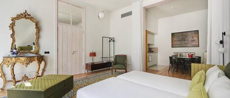 A hotel room with a large bed, a green ottoman, a mirror with an ornate frame, a desk with a lamp, and a dining area visible through an open door.
