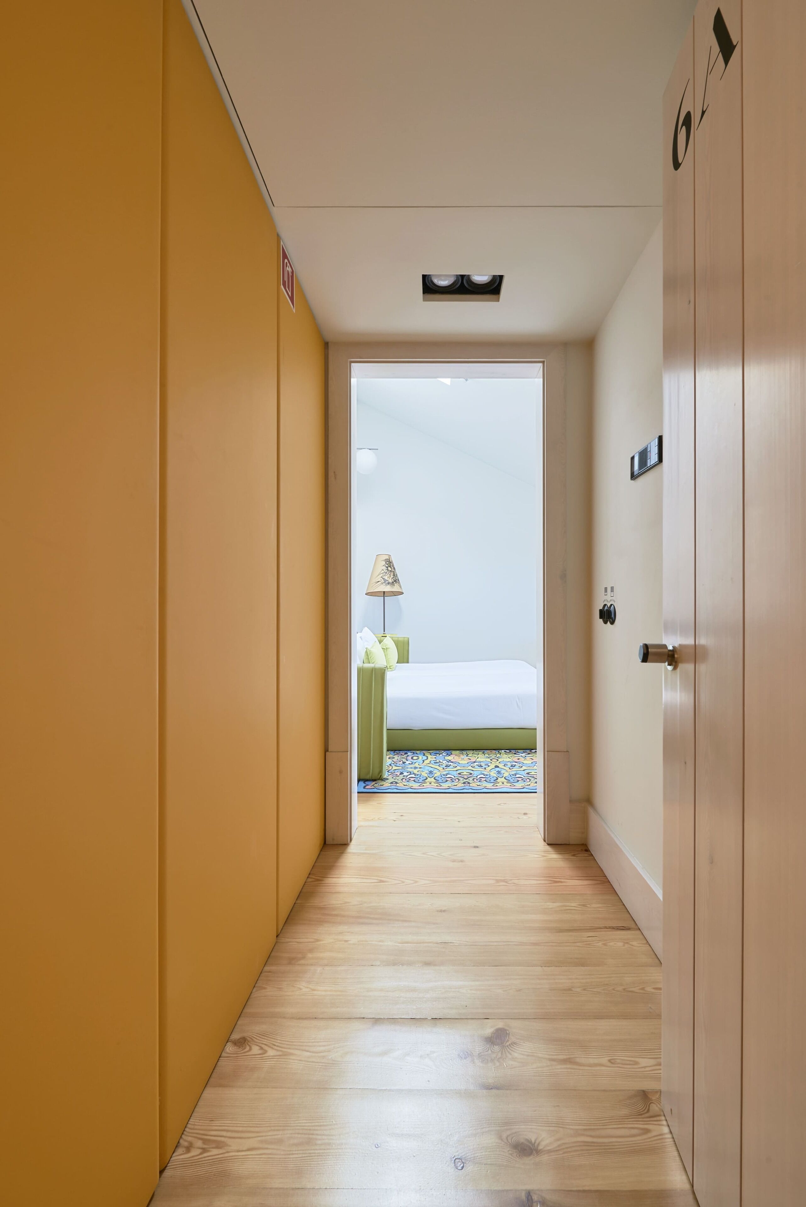A narrow hallway with wooden flooring leading to a room with a bed and a lamp.