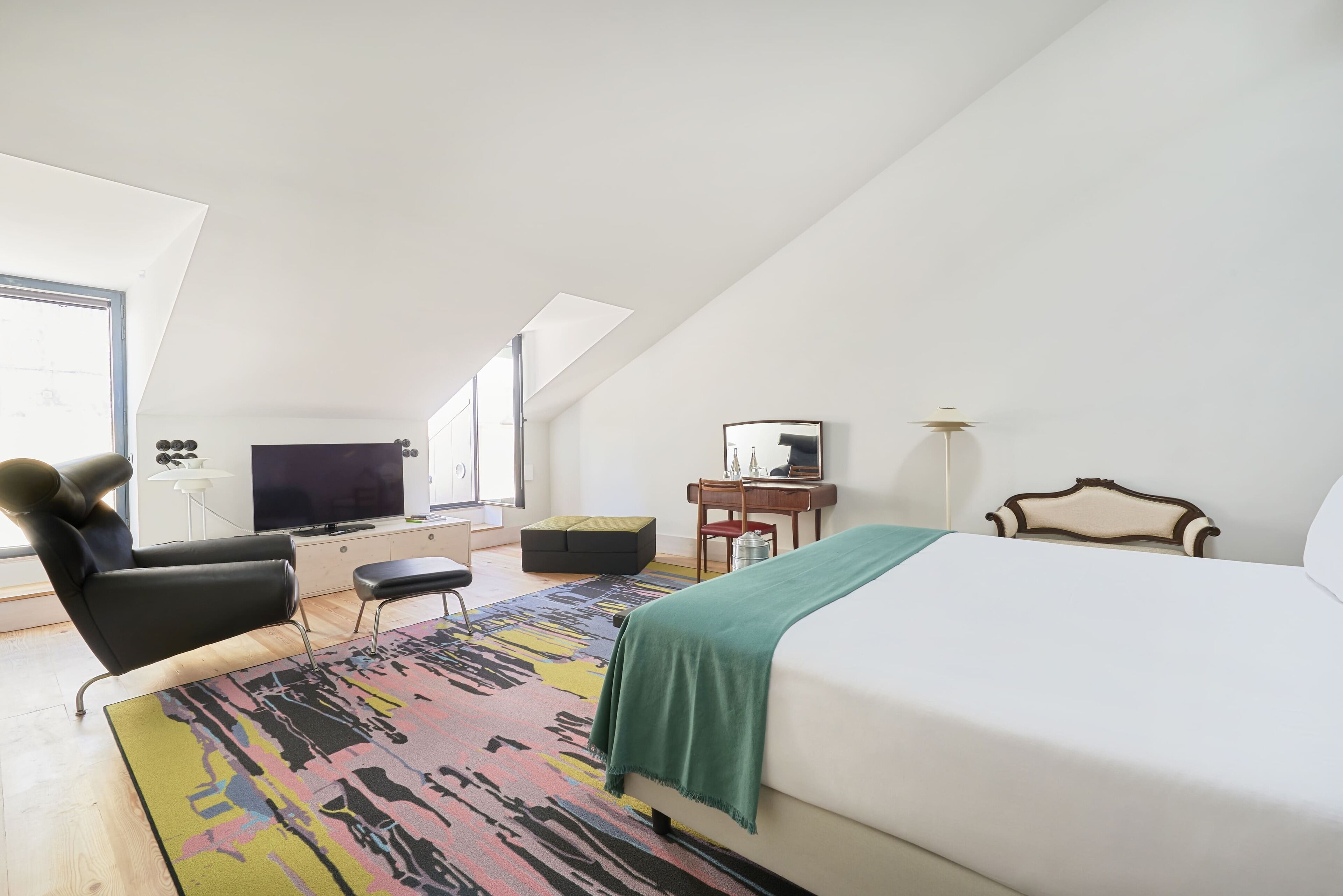 A modern hotel room with a large bed, a desk with a chair, a television, and a window providing natural light.
