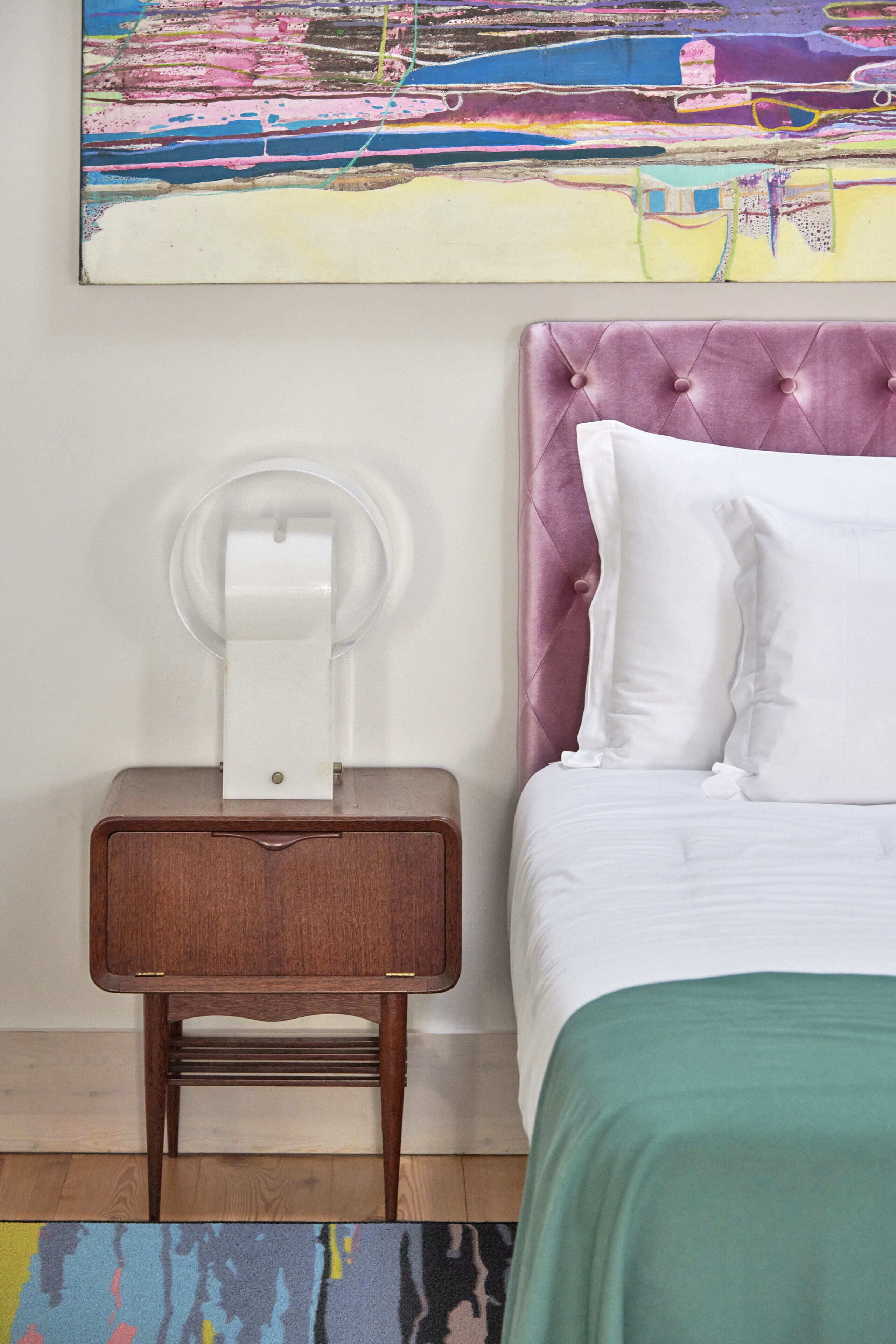 A neatly made bed with white linens and a green throw, a wooden nightstand with a white lamp, and a colorful abstract painting on the wall.