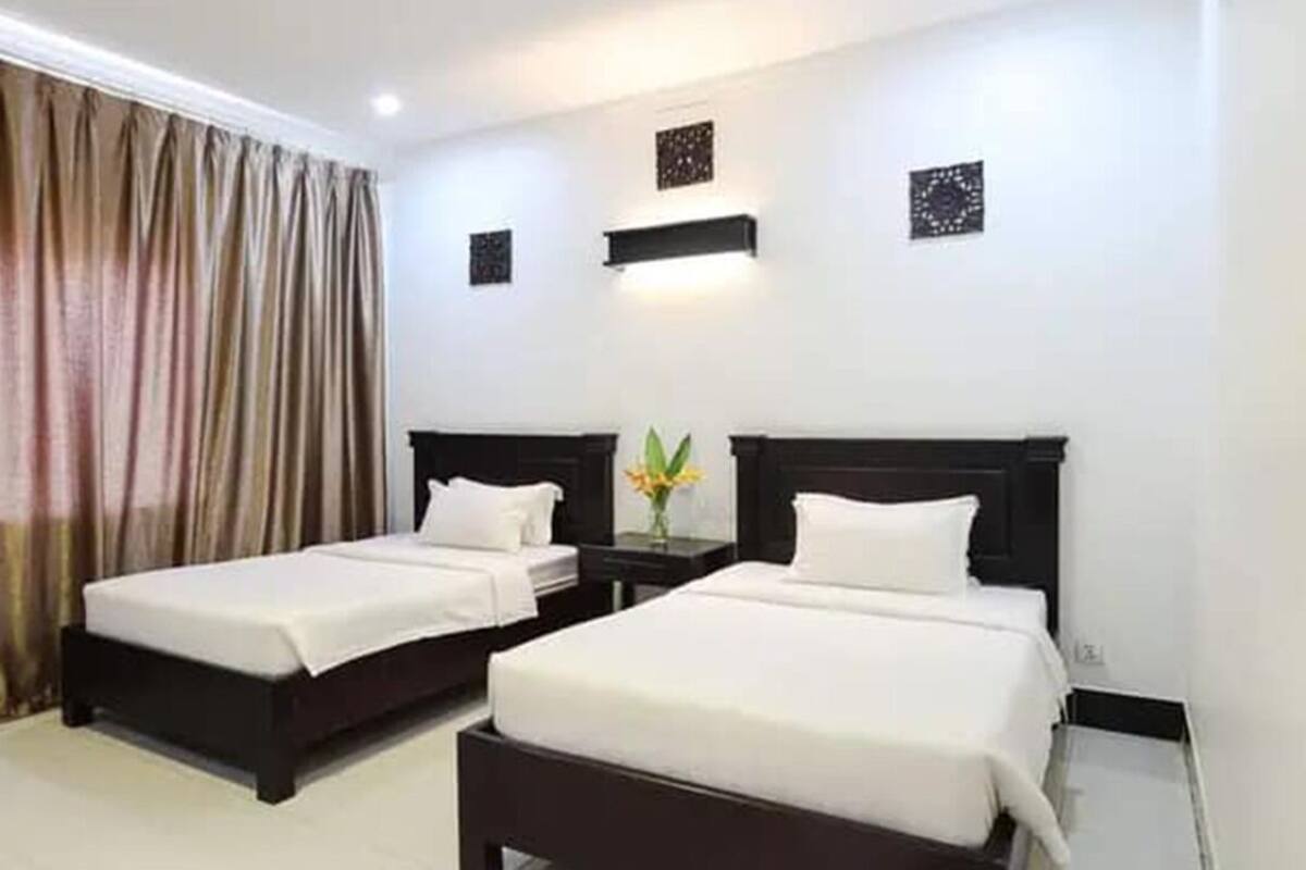 Quarto de hotel com duas camas de solteiro, uma mesa de cabeceira com abajur e uma janela grande com cortinas.