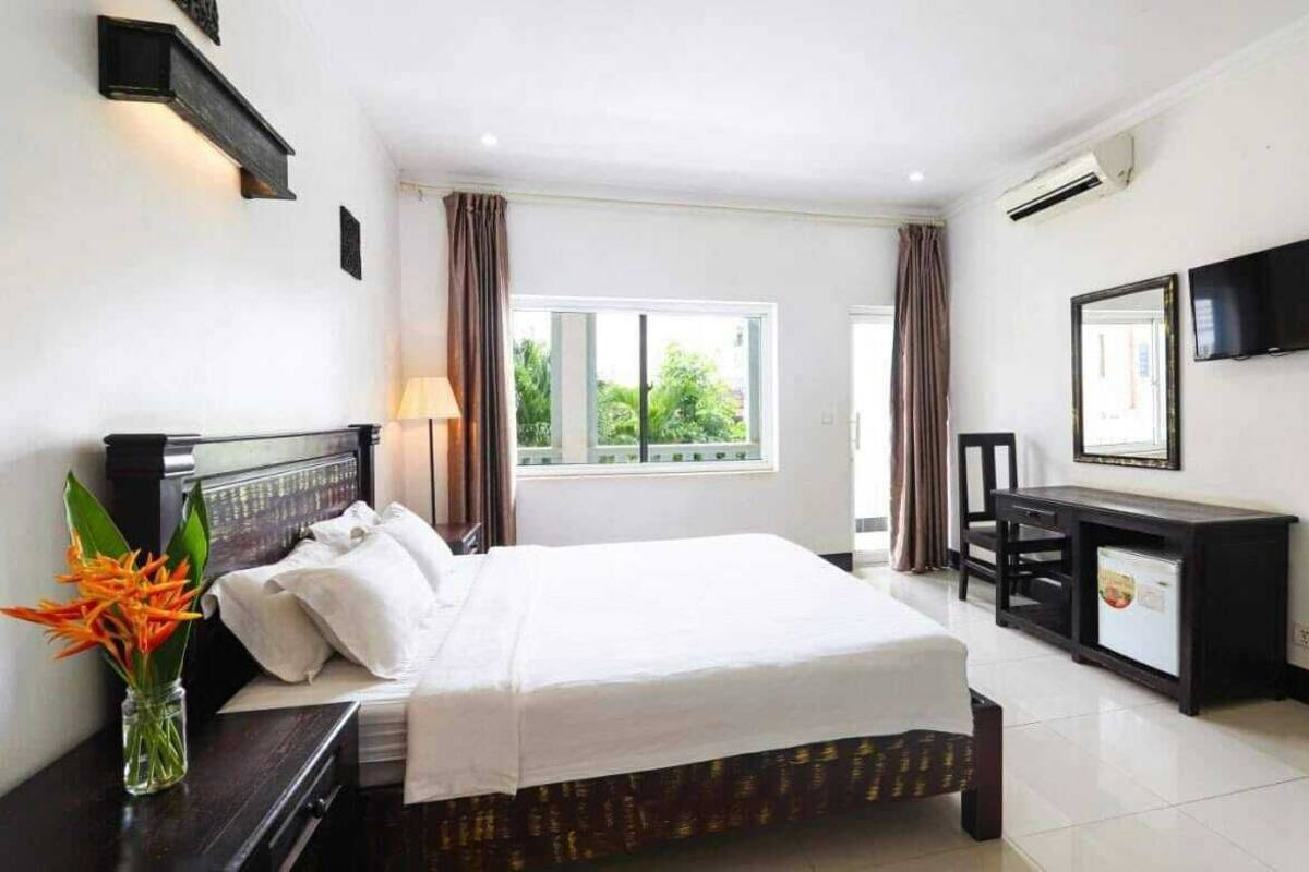 Quarto de hotel com cama, mesa de cabeceira, luminária, televisão e escrivaninha com computador.