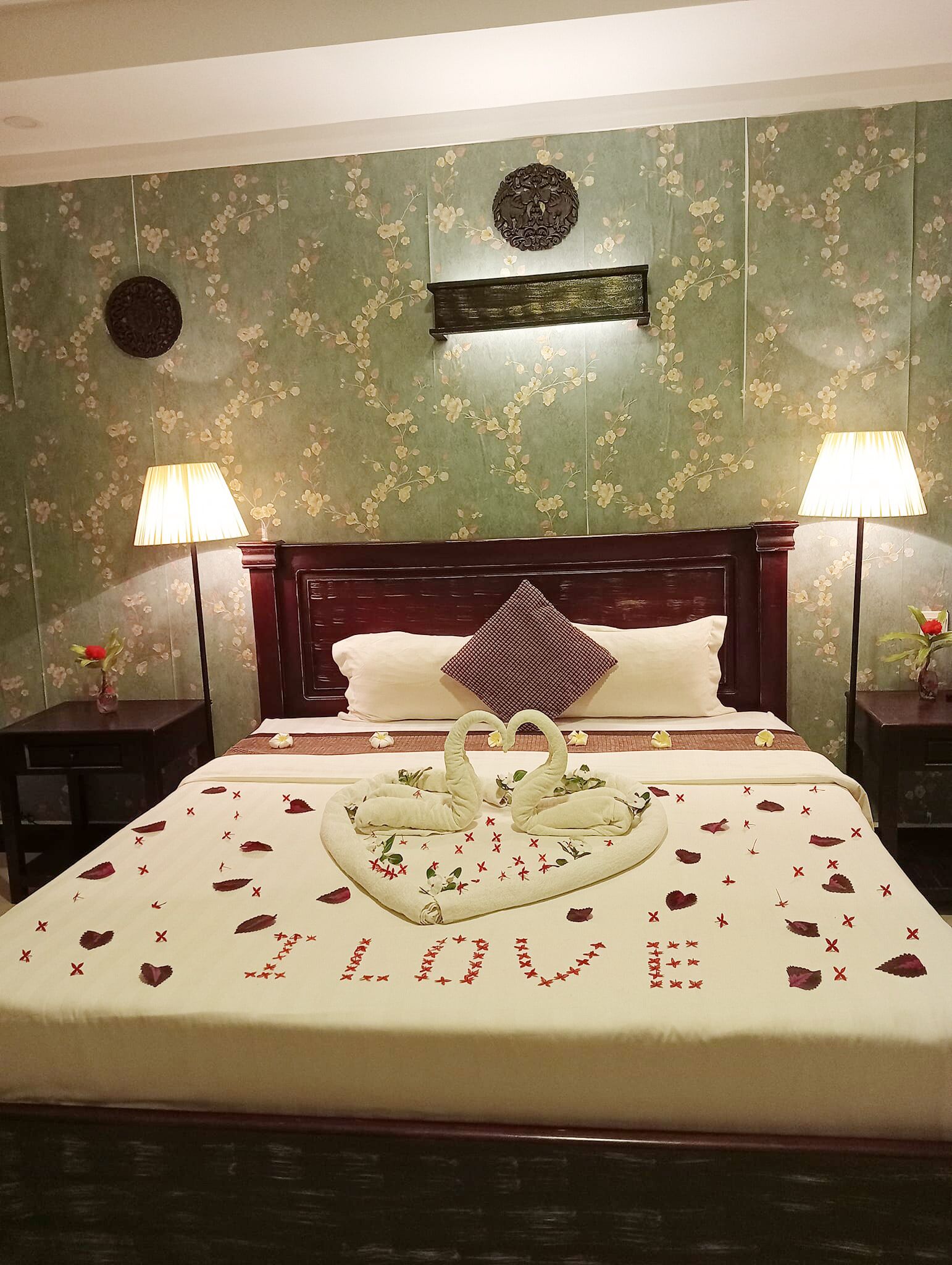Ett hotellrum med en stor säng, blommig tapet, två sänglampor och en dekorativ svan som mittpunkt på bordet.