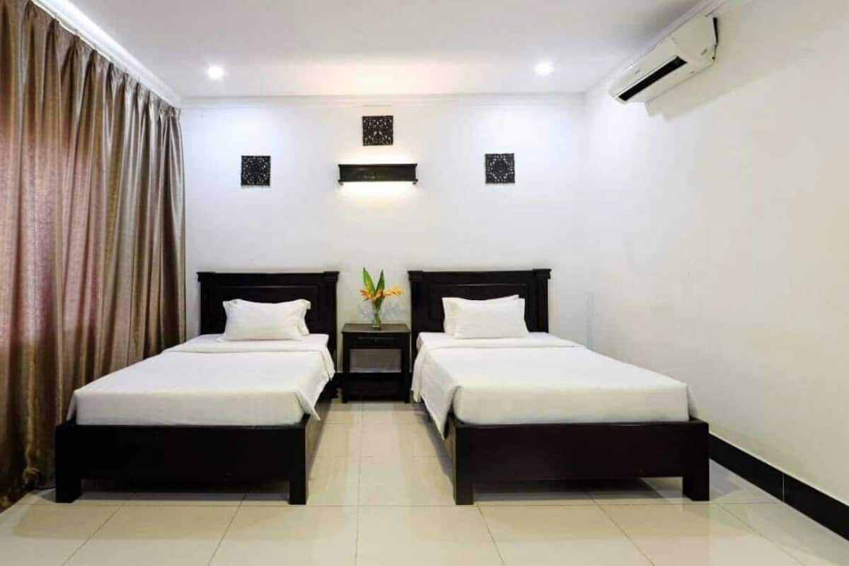 Quarto de hotel com duas camas de solteiro, uma mesa de cabeceira entre elas, um vaso com flores e um aparelho de ar condicionado na parede.
