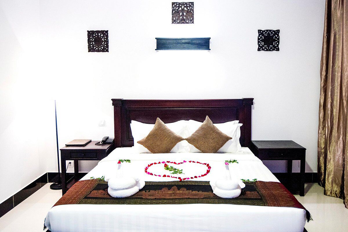 Una cama bien hecha con una decoración en forma de corazón, dos almohadas, una mesita de noche con un teléfono y una pared con paneles decorativos.