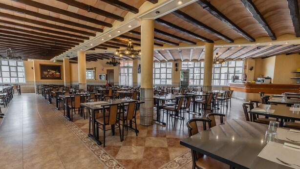 Un restaurante espacioso con techos altos, vigas de madera y ventanas amplias. Las mesas están ordenadas con sillas, y se ve una barra de fondo.