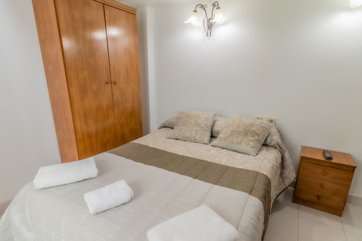 A bedroom with a bed, two pillows, a bedside table with a remote, and a wardrobe.