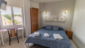 Un dormitorio con cama, escritorio y silla. Se ve una ventana con vistas a edificios y árboles.