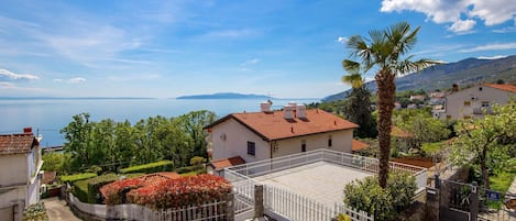 Zona residencial costera con una casa de techo de tejas rojas, un balcón con barandilla blanca y vista al mar.
