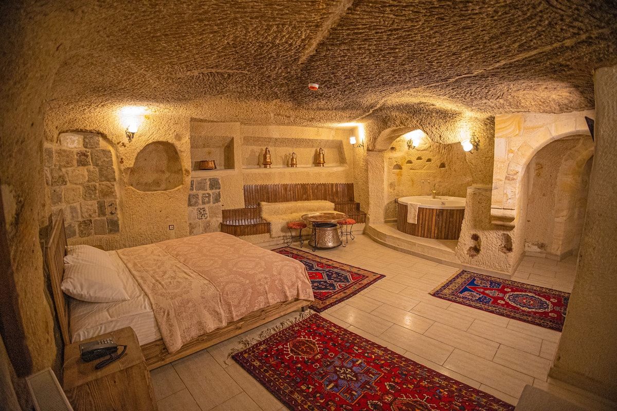 Un dormitorio con paredes de piedra, una cama, una mesita y una mesita de noche. Hay un baño con lavamanos y espejo.