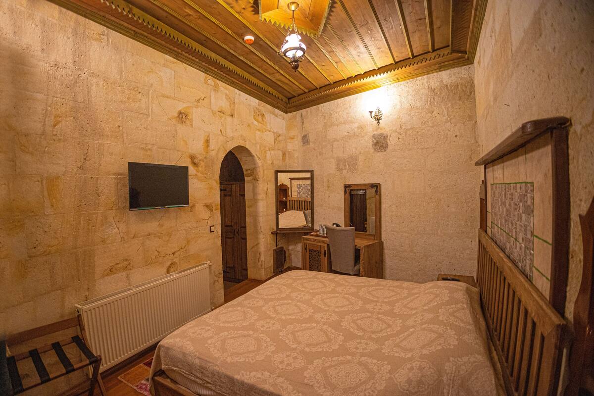Un dormitorio con paredes de piedra, techo de madera, cama con colcha estampada, escritorio con silla, espejo y televisor en la pared.