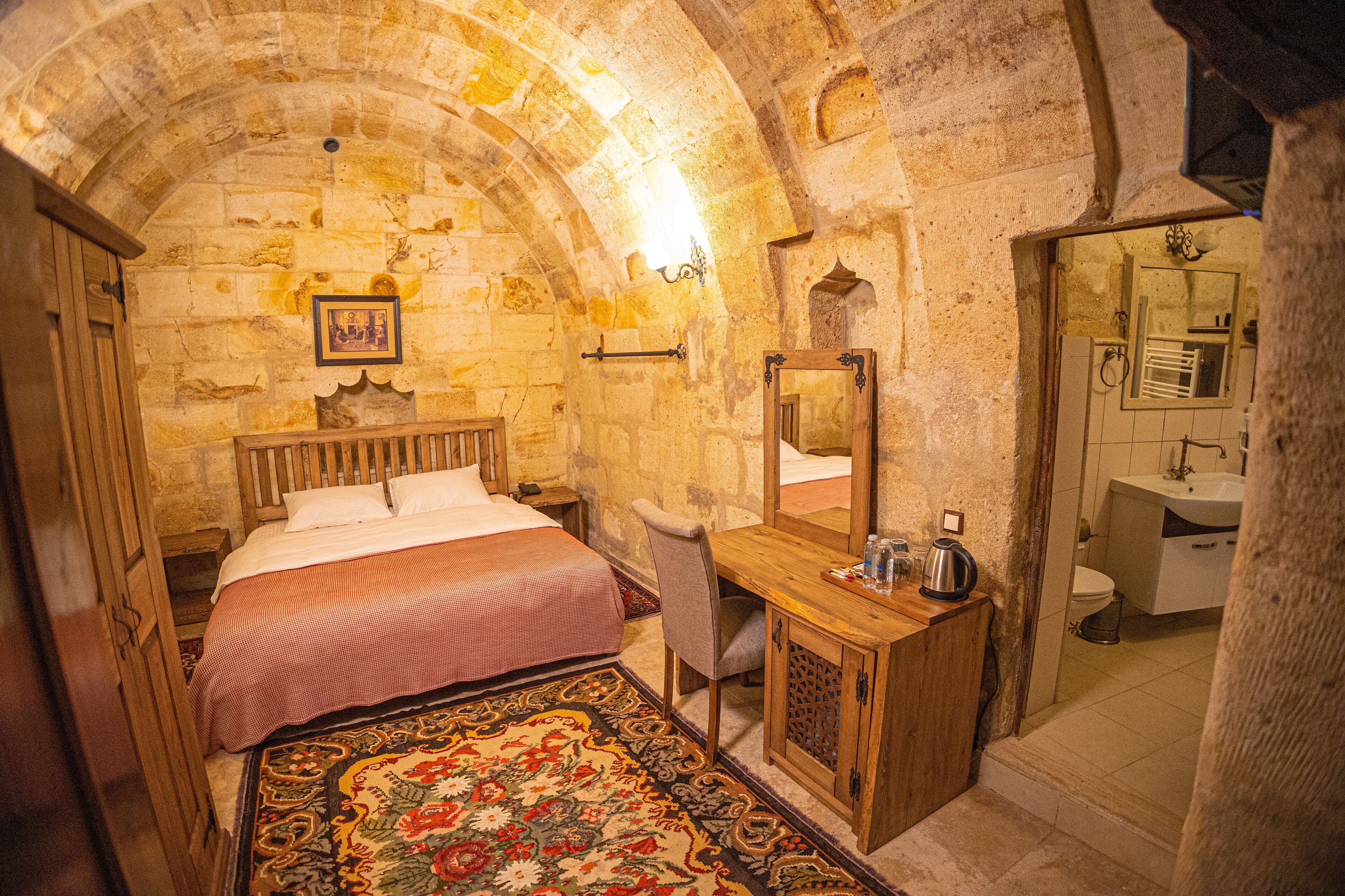 Una stanza con un letto, una scrivania, una sedia, uno specchio e un bagno visibile tramite un arco.