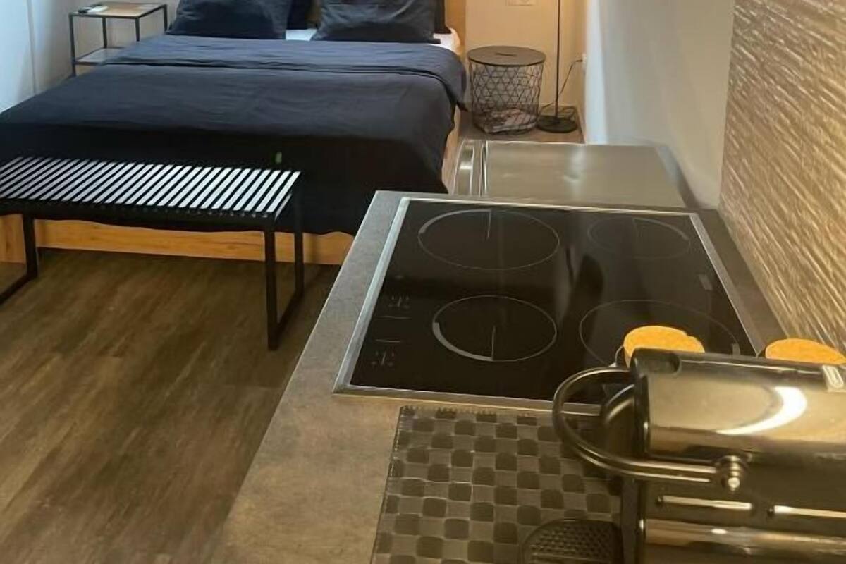 Une chambre compacte comprenant un lit, une table de chevet et un petit coin cuisine équipé d’une cuisinière et d’un évier.