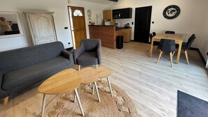 Un salón moderno con un sofá gris, una mesa de centro de madera y un sillón gris. Hay una zona de cocina con un refrigerador negro y una encimera de madera.