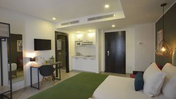 A modern hotel room with a bed, a desk with a chair, a kitchenette, and a bathroom.