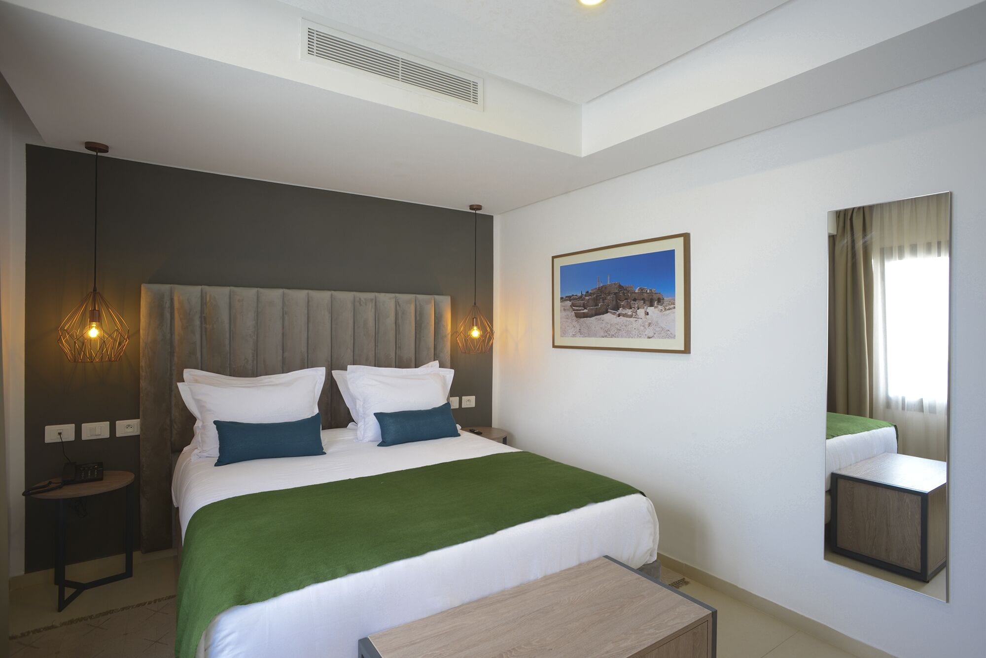 A modern hotel room with a large bed, a bedside table, a mirror, and a picture on the wall.