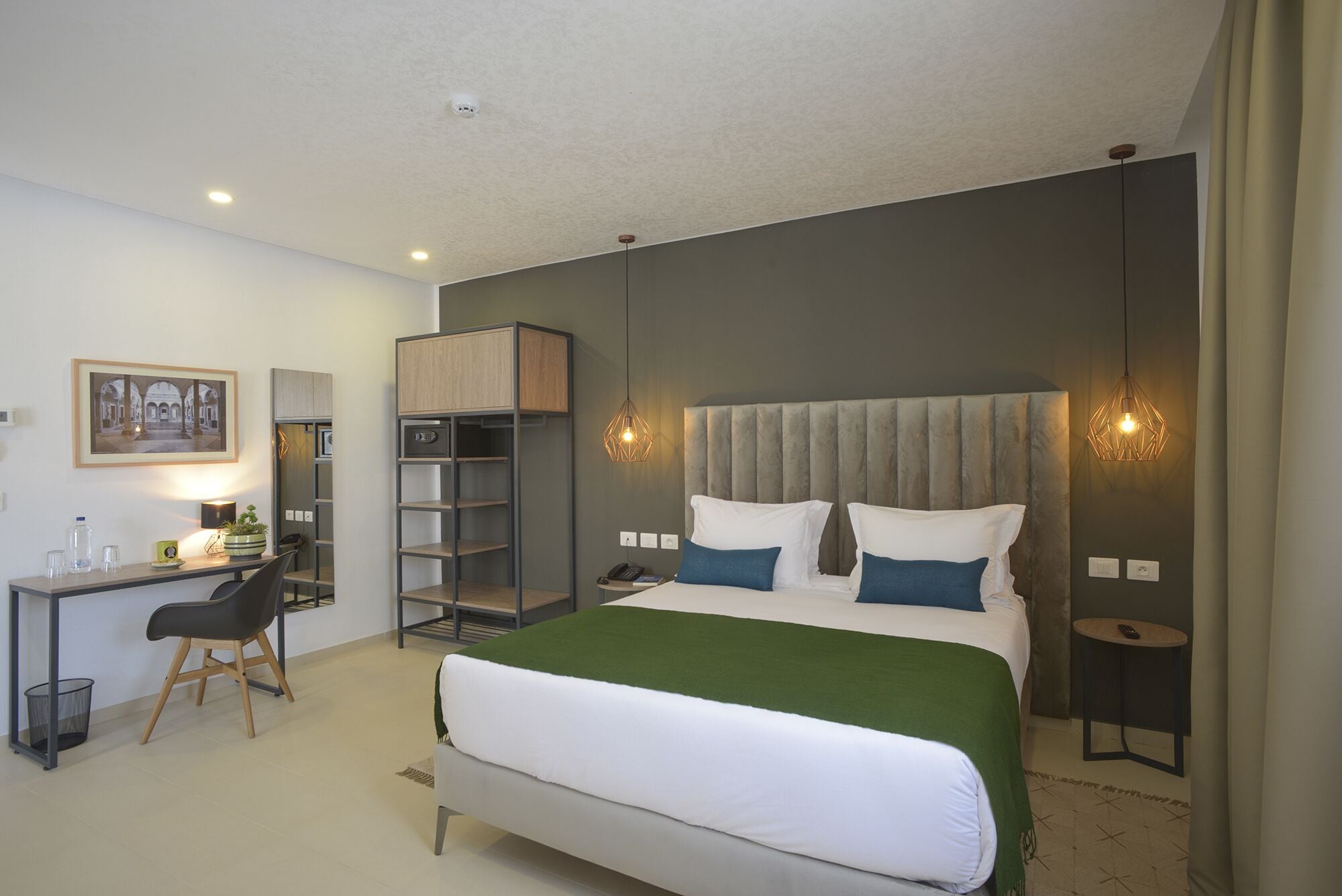 A hotel room with a bed, a desk with a chair, a bookshelf, and a small table with a lamp.