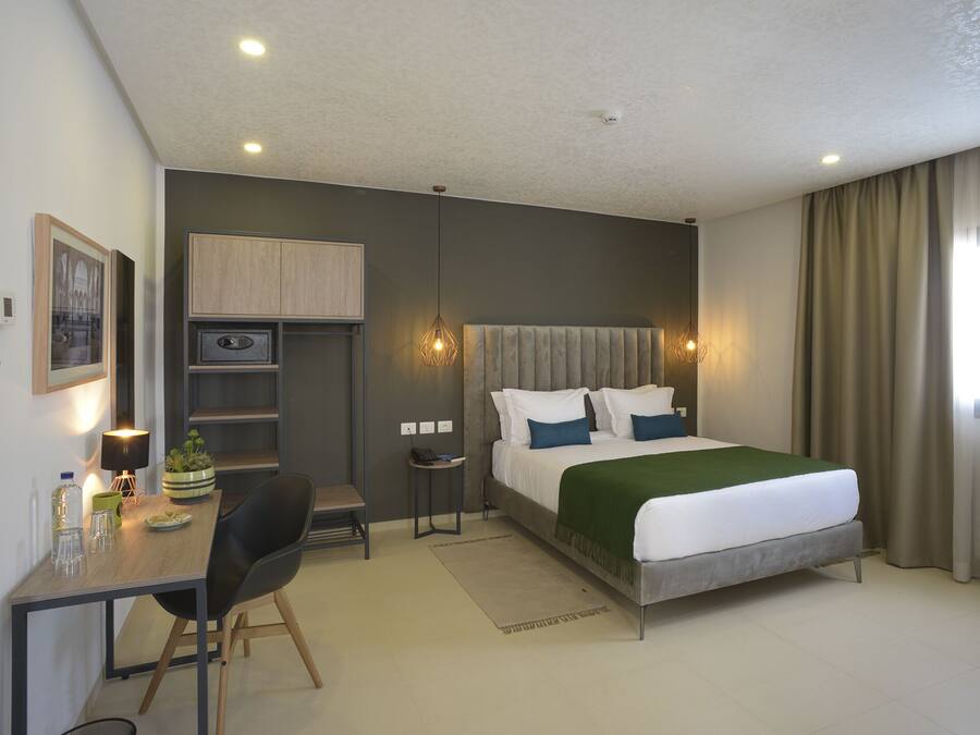 A modern hotel room with a bed, desk, chair, and a small kitchenette.