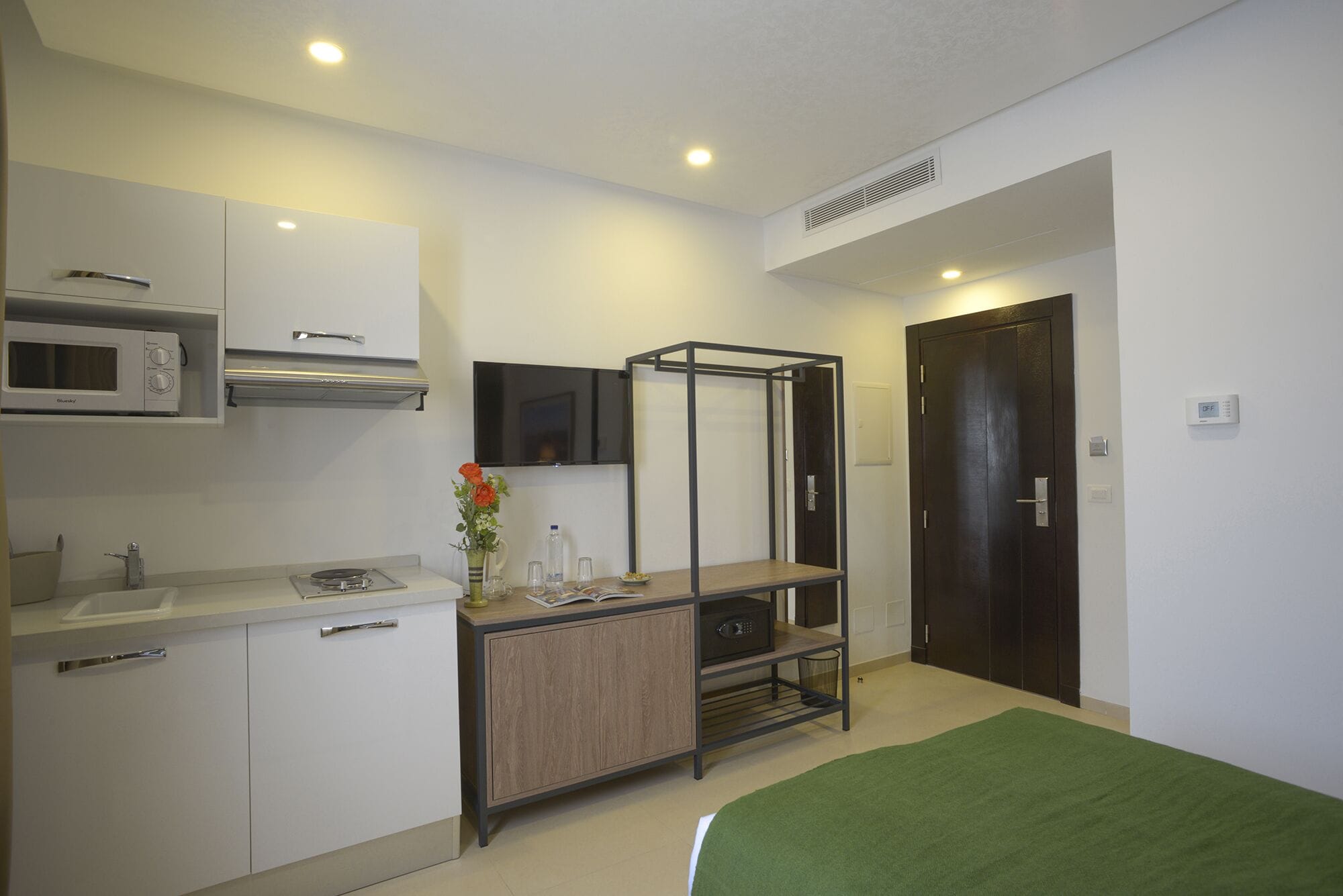 A compact hotel room with a kitchenette, a bed, a TV, and a dark wooden door.
