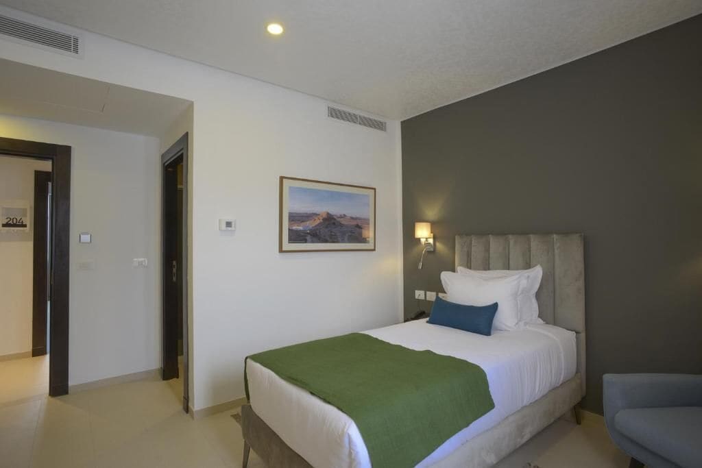 A hotel room with a bed, a green throw, a nightstand with a lamp, and a painting on the wall.