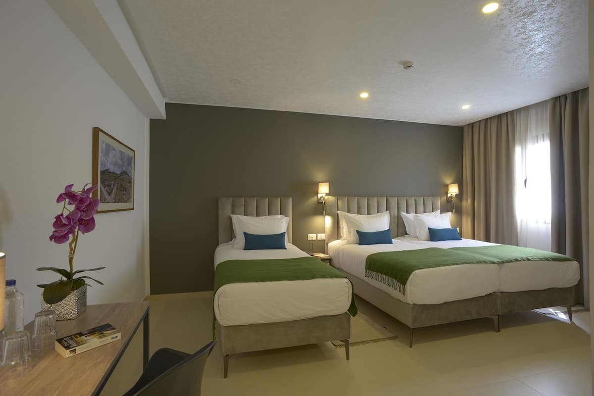 Une chambre d’hôtel avec deux lits, un bureau, une chaise, un vase avec des fleurs et un tableau au mur.