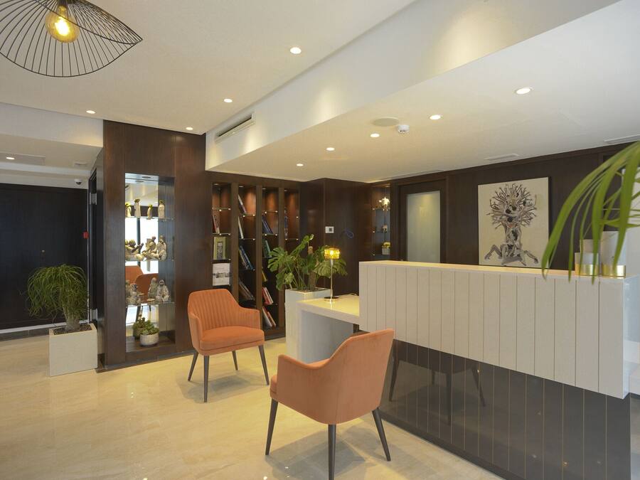 A modern reception area with a reception desk, two orange chairs, a large potted plant, and a wall-mounted artwork.