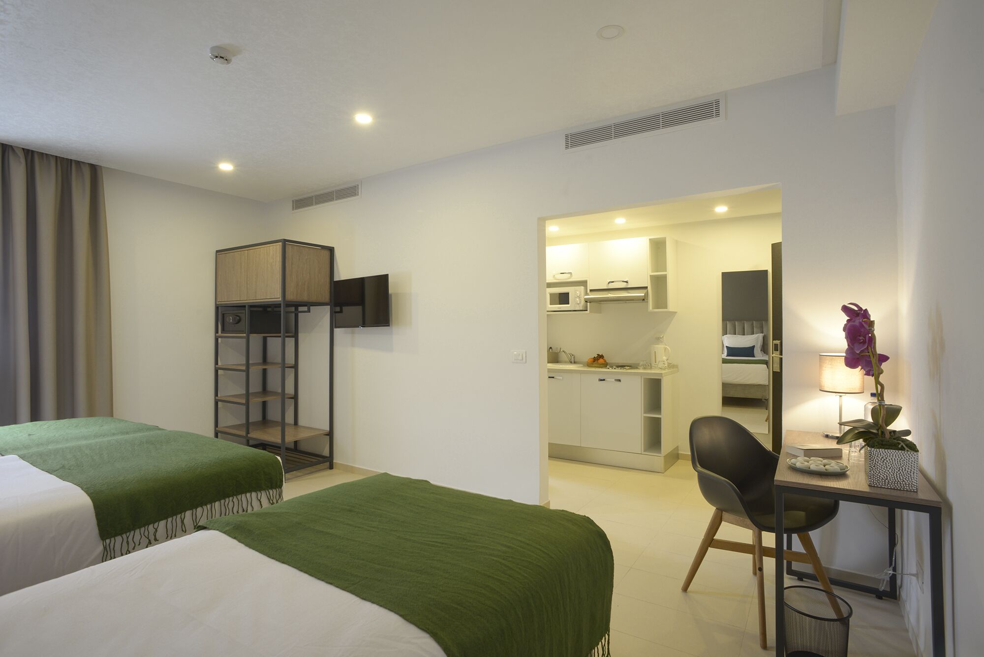 A hotel room with two beds, a desk with a chair, a kitchenette, and a TV mounted on the wall.