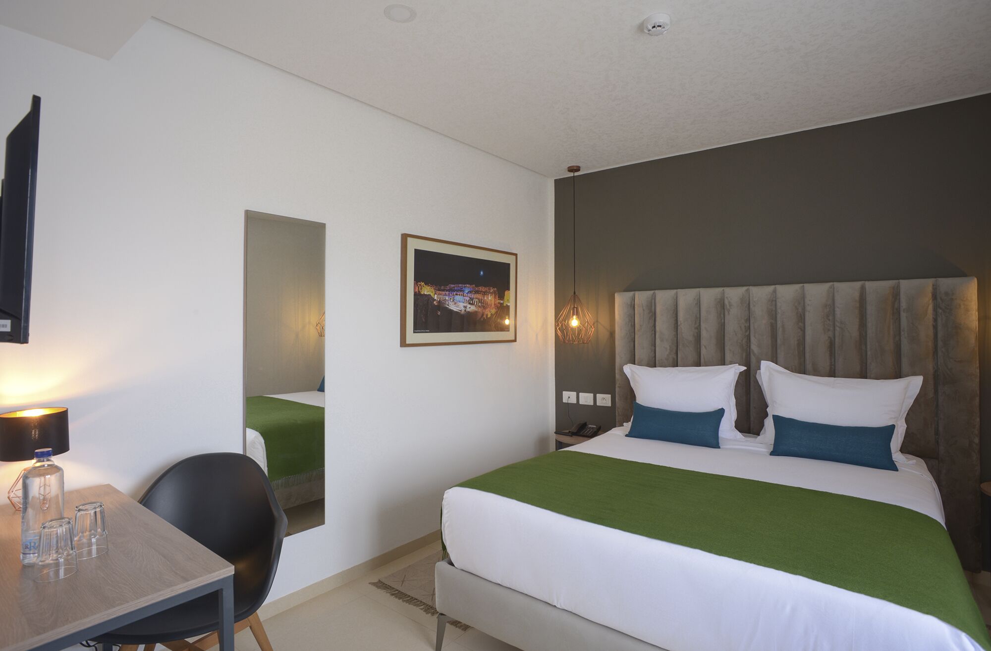 A hotel room with a bed, a desk, a chair, a lamp, and a framed picture on the wall.