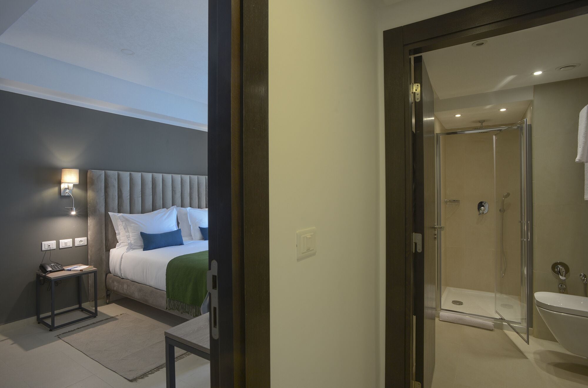 A bedroom with a bed, bedside table, and a nightstand. There is a bathroom with a shower, sink, and toilet.