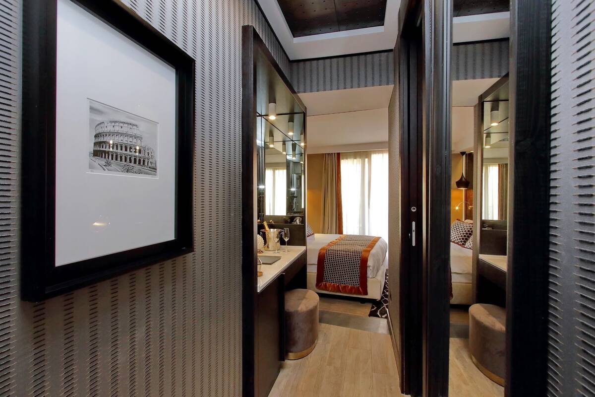 Una habitación de hotel con un cuadro enmarcado del Coliseo, una cama con una colcha estampada, un taburete pequeño, un escritorio con una lámpara y un espejo.