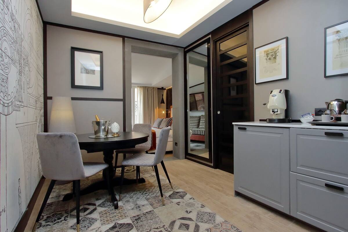 Un comedor moderno con una mesa redonda, sillas y un tapete estampado. Se observa una zona de cocina con armarios y una máquina de café.