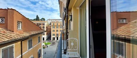 A narrow balcony with a metal chair, a glass door, and a view of a street with buildings and a clear sky.