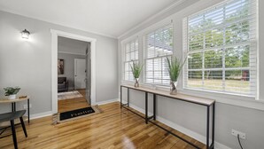 Un salón moderno con suelo de madera, una consola con plantas en macetas y ventanas amplias con persianas.