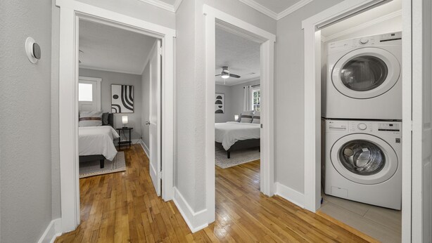 Un lavadero con lavadora y secadora de carga frontal, suelo de madera y un pasillo que conduce a un dormitorio con cama y mesita de noche.