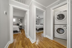 A laundry room with a front-loading washer and dryer, wooden flooring, and a hallway leading to a bedroom with a bed and nightstand. - Cozy 2BR Cottage in Downtown Warm Springs (A) (Warm Springs)