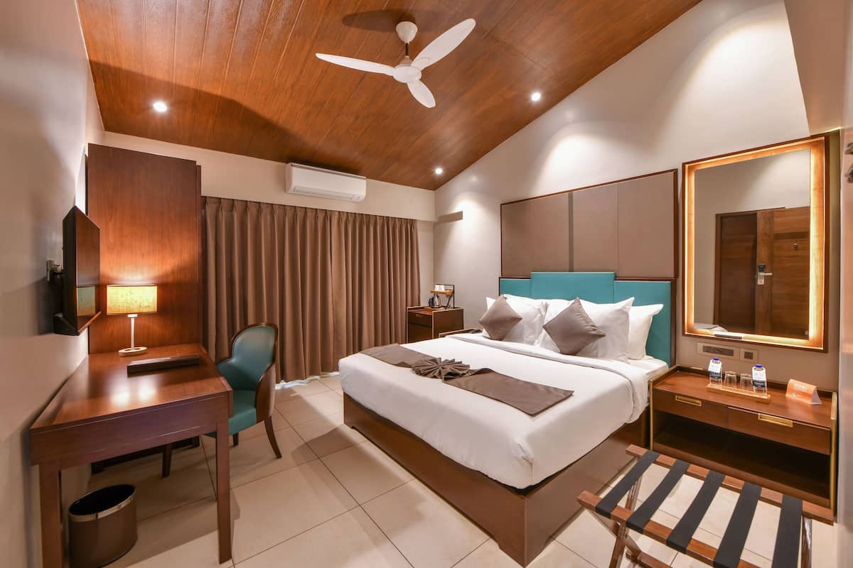 A modern hotel room with a wooden ceiling, a bed with white bedding, a desk with a lamp, a chair, a bedside table with a mirror, and an air conditioning unit.