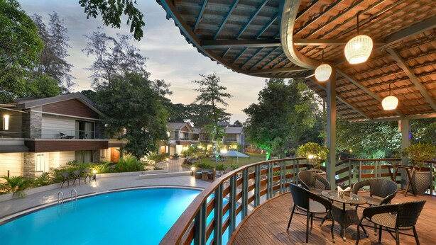 A swimming pool with a wooden deck and wicker furniture, surrounded by houses and trees. - Arova Woods Zinc Journey by The Fern (Mawal)