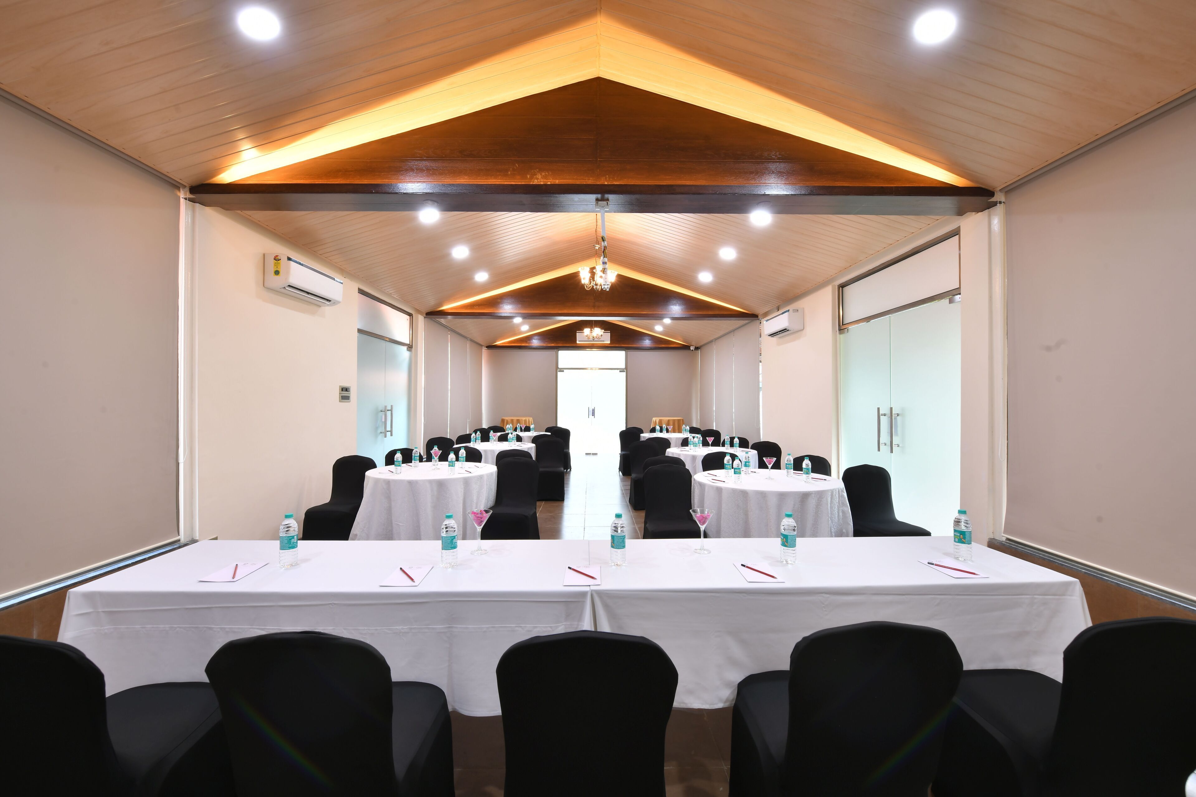Une salle de conférence avec plusieurs tables, des chaises noires, des nappes blanches, des bouteilles d’eau et un lustre.