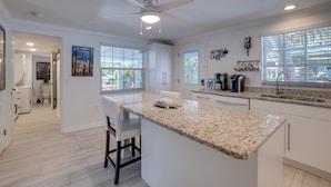 A modern kitchen with white cabinetry, granite countertops, and a central island. There is a ceiling fan, a window with blinds, and a coffee machine on the counter. - Private Pool - Anna Maria Hideaway by SeaBreeze Vacation (Holmes Beach)