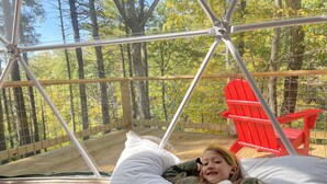 A cozy outdoor sleeping area with a metal frame, a red chair, and a bed with pillows. - Seasonal, Cozy Geo-Dome with Heat/AC on Private Hiking paradise in Gunks. (Wallkill)