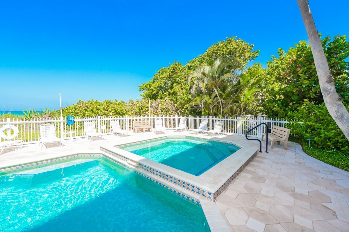 A pool area with two rectangular pools, surrounded by a white fence, benches, and palm trees.