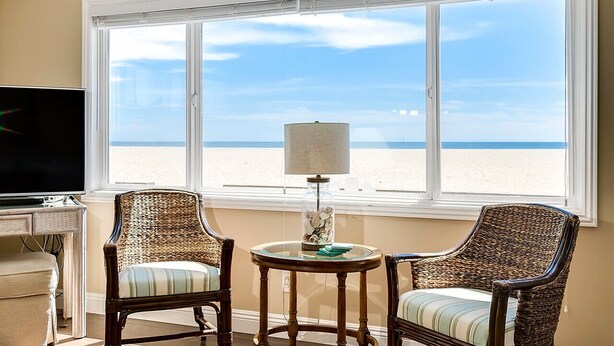 Habitación con un amplio ventanal con vista a la playa, dos sillones de mimbre, una mesita con una lámpara y un televisor sobre un mueble.