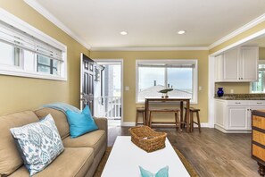 A living room with a beige sofa, a white coffee table, and a kitchen area with white cabinets and a dining table.