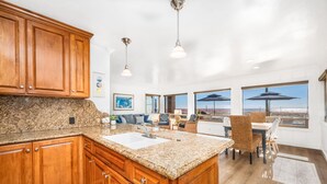 A kitchen with wooden cabinets, granite countertops, a central island, pendant lighting, and large windows overlooking a beach.