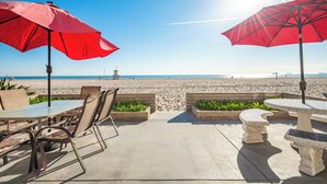 Un patio frente a la playa con mesas y sillas, sombrillas rojas y vista al océano.