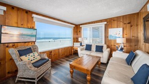 Una habitación acogedora con un gran ventanal con vistas a la playa, una silla de mimbre, un sofá blanco y una mesa de centro de madera.