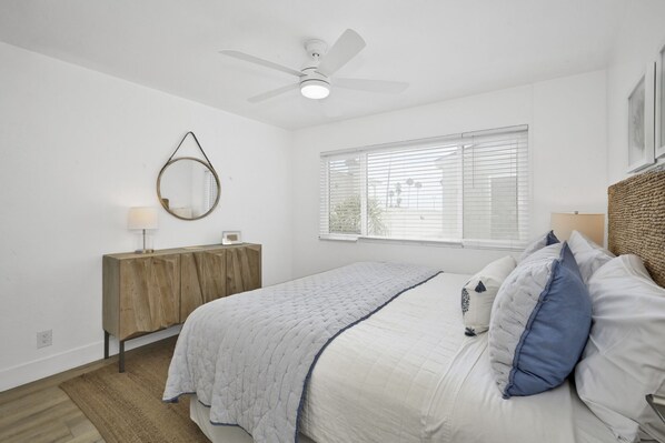 A bedroom with a bed, a wooden dresser, a round mirror, a ceiling fan, and a window with blinds.