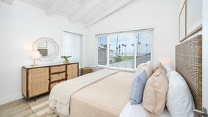 Een slaapkamer met een groot bed, een rieten dressoir, een spiegel en uitzicht op palmbomen door het raam.