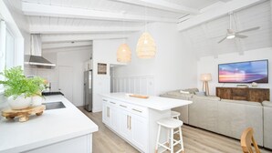 Een moderne keuken met witte kasten, een centraal eiland, hanglampen en een zithoek met een bank en een televisie.